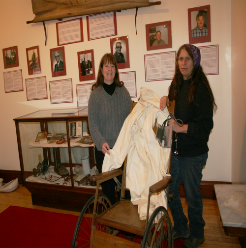 Gail Murray, left, Historical Association Executive Director, and volunteer, Deb Evans, stand next to an early wooden wheelchair, while holding up a physician's coat worn by Dr. Stuart Nelson. Many other artifacts will be on display in the Medicine Up North Exhibit that opens Friday, November 25. Photo by Carol Hansen.